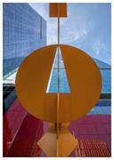 Large orange geometric sculpture in front of reflective modern glass buildings, viewed from below.
