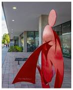 Red abstract metal sculpture of a human figure stands on a brick sidewalk near a modern building with glass windows.