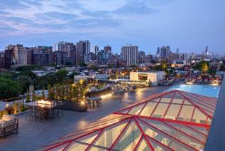 Rooftop view with pool, city skyline, outdoor seating, and glass roof at dusk.
