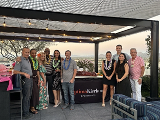 A group of people wearing leis pose for a photo at an outdoor event with city views in the background.