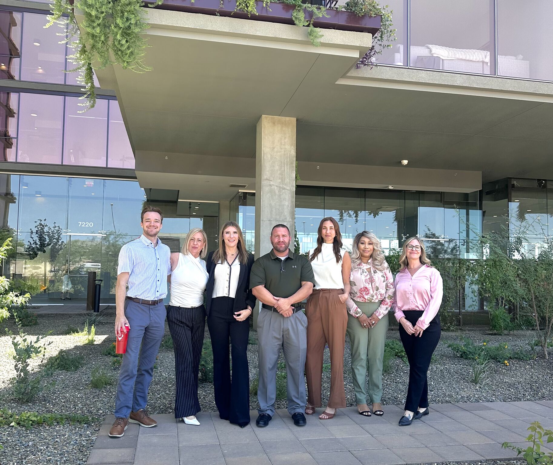 Seven people stand smiling in a row outside a modern building, celebrating their years with the Optima Community and enjoying vibrant greenery.
