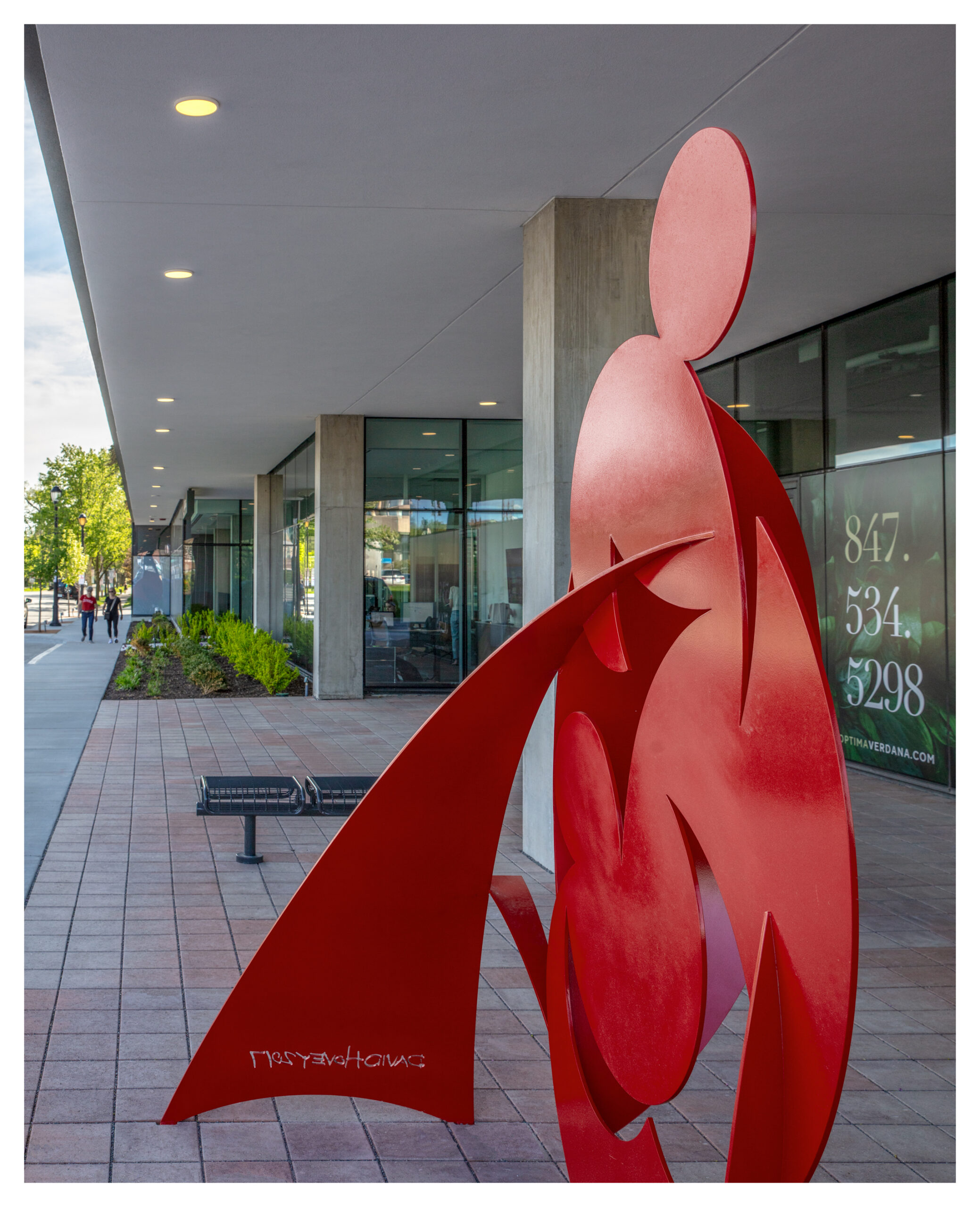 Red abstract sculpture outside a modern building with glass windows reflects vibrant Optima Community living. Call 847-534-5298.