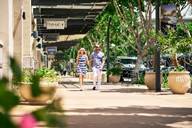 Two people walk on a sunny sidewalk lined with shops, trees, and large potted plants.