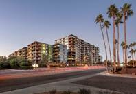Modern apartment buildings with balconies at dusk, palm trees in the foreground, and blurred car lights on the street.