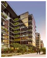 Modern apartment building with glass walls, balconies, and greenery along a sidewalk at sunset.
