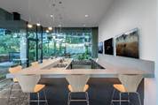 Modern kitchen with a light wood island, four yellow chairs, large windows, and wall-mounted TVs.