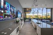 Modern kitchen with large windows, multiple wall-mounted TVs, and a long island with a sink and pendant lights.