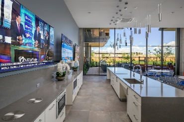 Modern kitchen with gray countertops, pendant lights, and wall-mounted TVs, overlooking a patio and lawn.