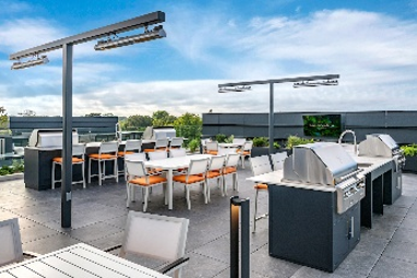 Modern outdoor rooftop patio with grills, dining tables, white chairs, and blue sky in the background.