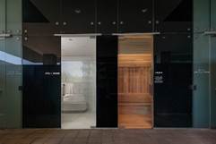 Three glass doors reveal a modern steam room on the left and a wooden sauna on the right inside a spa facility.