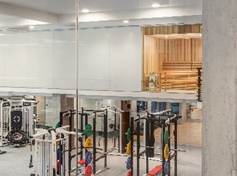 Modern gym with weight machines and benches, viewed through glass; a sauna room with wooden benches is visible in the background.