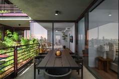 Modern balcony with a wooden dining table, chairs, railing with plants, and city view through glass doors.