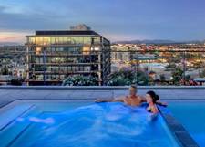 A couple relaxes in a rooftop pool at dusk, with city lights and a modern building in the background.