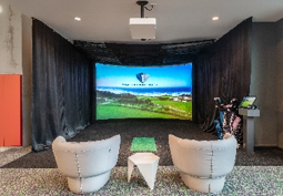 Two chairs face a curved golf simulator screen, with golf clubs and a touchscreen nearby in a modern room.