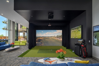 An indoor golf simulator room featuring a large screen, green turf, black curtains, a golf bag, and lounge seating with blue couches.