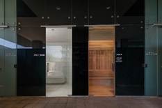 Three glass doors; the left leads to a white room, the center to a dark room, and the right to a warmly lit wooden sauna.