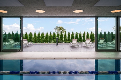 Indoor pool with blue water, looking out to an Optima patio area with lounge chairs and green shrubs under a blue sky.