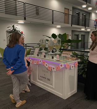 Three women stand by a decorated refreshment table with drinks and cups in a modern building lobby.