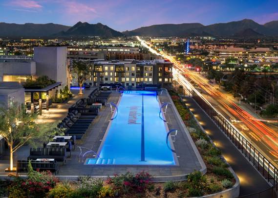 Rooftop pool lit at dusk, with city lights, mountains in the background, and traffic on a busy road beside the building.