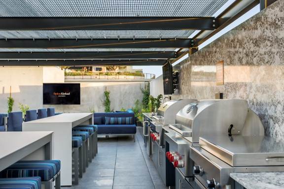 Modern outdoor kitchen with stainless steel grills, a dining table, blue cushioned seating, and a TV on the wall.