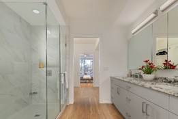 Modern bathroom with a glass shower, gray vanity, granite countertop, and red flowers, leading to a bedroom.