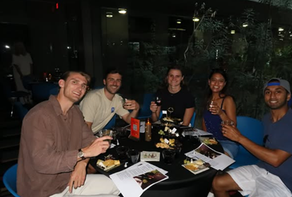 Five people sit around a round table, smiling and raising glasses, with plates of dessert and papers in front of them.