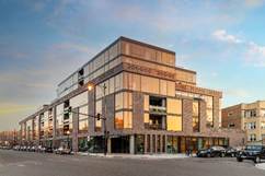 Modern multi-story building with large windows on a city corner, reflecting the sunset sky, with cars parked along the street.