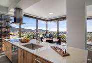 Modern kitchen with large windows, mountain views, and a dining area. Countertops have a sink, fruit bowl, and tray.